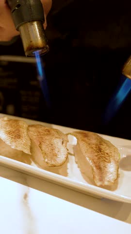 A close-up shot shows a chef searing nigiri sushi with a blue blowtorch flame