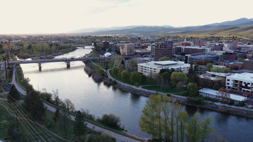 Missoula Montana downtown skyline along Clark Fork River at sunset, riverside park with walking path, historic bridge, brick buildings and bare rolling hills under soft pastel sky. Aerial wide shot.