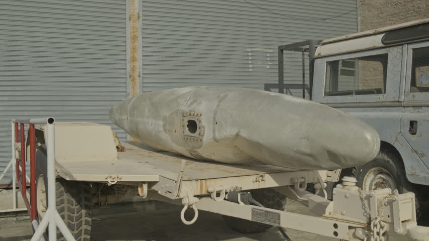 A side view of a large metal fuselage or aircraft part loaded onto a rugged military trailer, showcasing heavy-duty transport gear in an outdoor facility.