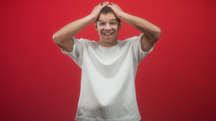 Man with fists raised and glasses, hands clenched near chest, wearing white t shirt in studio red backdrop; playful joy.