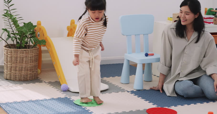 Happy Asian little girl jumping on colorful sensory stepping stones mats with her mother cheering nearby.