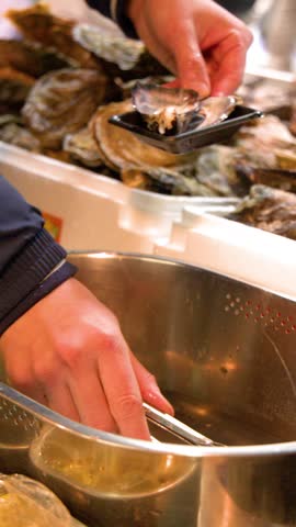 A vendor uses metal tongs to place fresh oysters into small trays at a market.