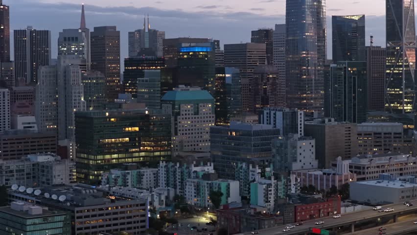 San Francisco California dense downtown skyline at dawn with illuminated glass towers and Transamerica Pyramid, mixed residential and commercial rooftops glowing under blue twilight sky. Aerial view.