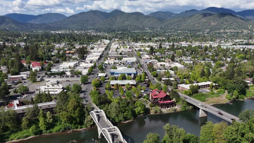 Grants Pass Oregon white arch bridge with truck crossing over Rogue River, red-roof historic building among dense green trees, low-rise downtown and forested mountain range backdrop. Aerial back wide