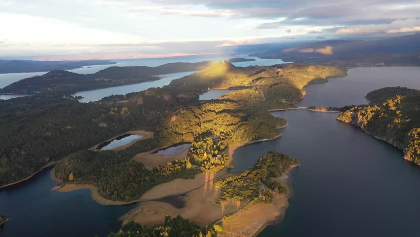Breathtaking drone panorama of the Bariloche lake district at sunset. Showcases the deep blue water, forested islands, and rolling mountain ridges bathed in warm golden light under a cloudy sky.