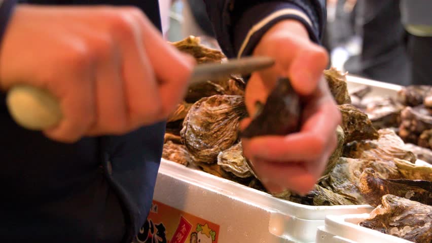 Close-up of hands shucking fresh oysters with a knife at a Japanese market.