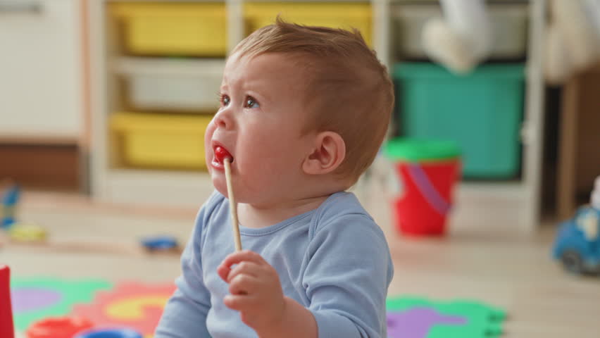 Toddler boy in blue shirt holds a red bead on a stick with his hand, touches his mouth, sits on a foam play mat amid toys and a red bucket in a home playroom building; curious exploration.