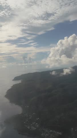 Captured from a window seat during a flight from Ambon to Jakarta, this stunning aerial view highlights the rugged coastline of an Indonesian island fading into the vast ocean. Below, the dark green, mountainous terrain meets the sea along a winding, narrow shore dotted with faint traces of coastal settlements. The water