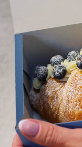 Displaying baked delicacy. Boutique bakery scene featuring crafted blueberry croissant presentation. Refined setting with styled pastry box featuring blueberry croissant and elegant manicure