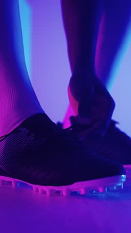 Close-up image of a football player tying the laces of cleats with vibrant purple and blue neon lighting emphasizing athletic footwear and preparation for a game