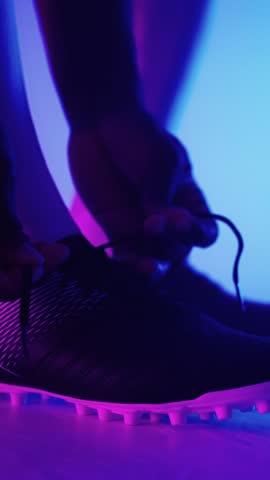 Close-up shots of a football player tying the laces of cleats under dramatic purple and blue lighting, highlighting preparation and focus before a match