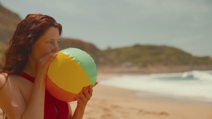 Woman with red hair prepares a colorful beach ball on the sandy shore, surrounded by waves and a bright summer sky