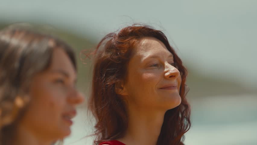 Two women at the beach, one in a red swimsuit and the other in a black and white bikini, enjoying the sun and ocean breeze with calm expressions