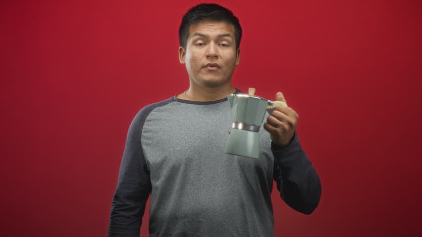Man holding a moka coffee maker by the handle while raising it and mouthing an exclamation against a red studio backdrop; surprise curiosity.