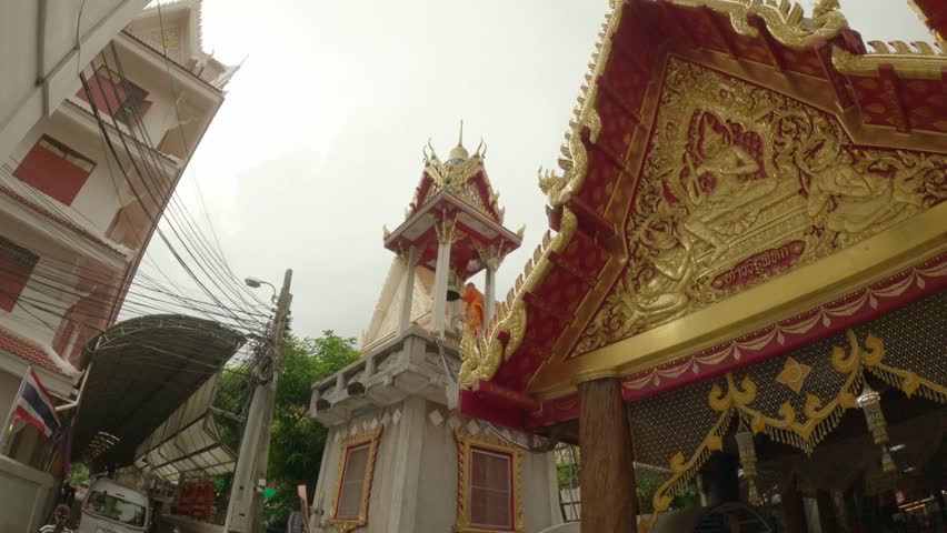 The video footage captures a scene within a temple complex, focusing on a traditional Thai bell tower (Ho Rakhang). A Buddhist monk dressed in bright orange robes is seen standing on the upper platform of the tower, systematically striking the large ceremonial bell. In the foreground, the ornate, golden pediment of a temple roof features intricate carvings of mythical figures, while a multi-story building and a web of overhead utility lines stand on the left under an overcast sky.