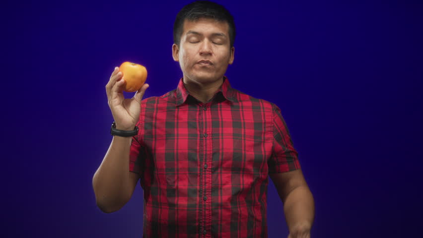 Man holding persimmon in right hand while left hand gestures near chest, wearing red plaid shirt against blue studio backdrop; thoughtful curiosity.