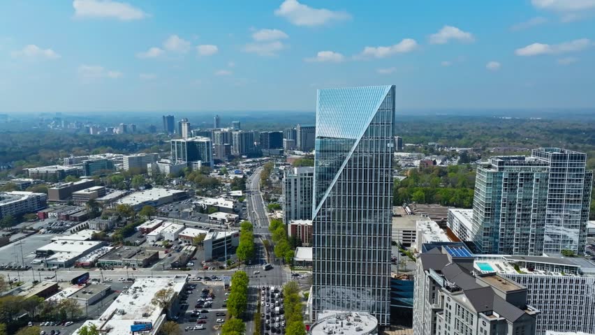 Atlanta modern glass skyscraper tower with diagonal facade, surrounding mixed-use buildings, freeway, tree-lined streets and distant downtown skyline under sunny sky. Georgia, USA. Aerial wide shot