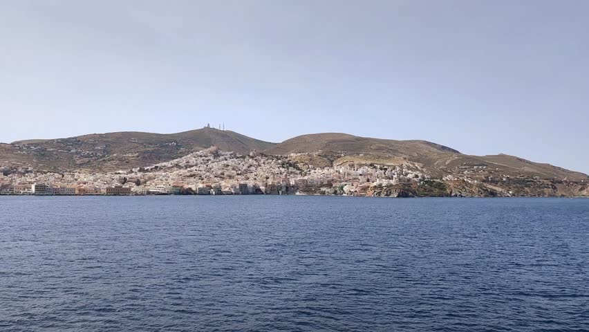 View of the vibrant port and waterfront promenade of Ermoupoli on Syros island, Greece, showcasing its colorful traditional neoclassical architecture and the historic hillside town of Ano Syros under a clear blue summer sky.
