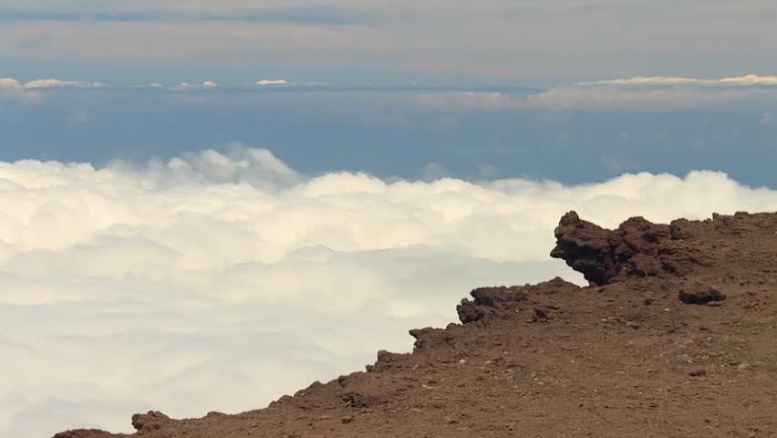 Sweeping static footage over the volcanic summit in Haleakala National Park, Maui, Hawaii, reveals a rugged rocky ridge above a thick blanket of rolling clouds beneath a tranquil blue sky.
