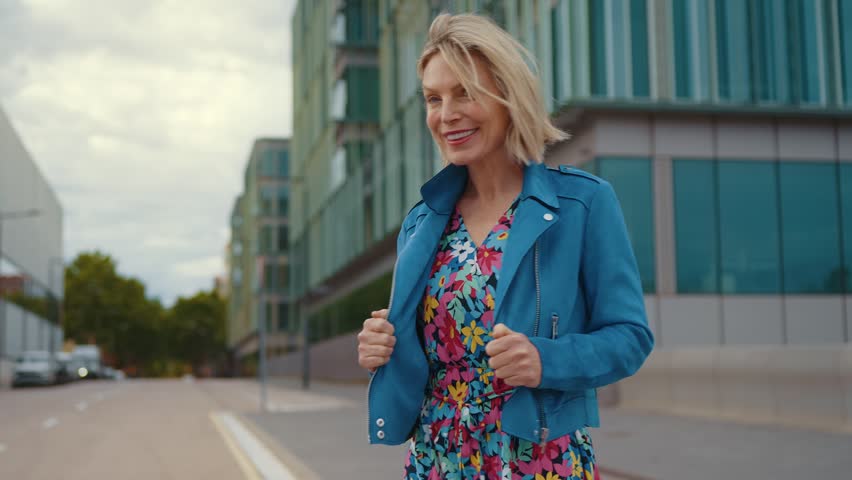 Elegant middle-aged woman with blonde hair enjoying a confident walk through a modern city, smiling with the wind blowing through her hair while wearing a stylish blue jacket and floral dress