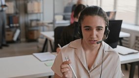 Handheld shot of young Caucasian support agent in headset providing help while sitting at desk in call center with colleagues in background - Powered by Shutterstock - Get 15% off with code: PIKWIZARD15