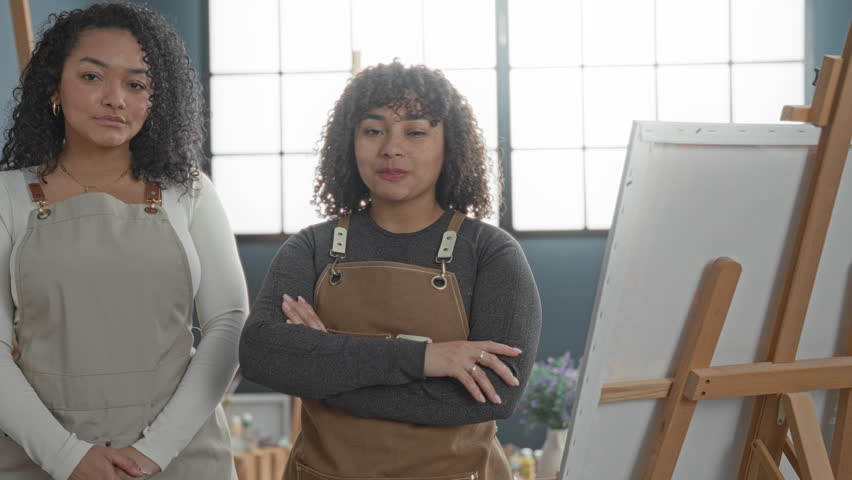 Woman artist wearing aprons, one with arms crossed and both smiling in studio; creative teamwork confidence.