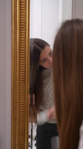Young girl with long brown hair is meticulously brushing her hair in front of gold-framed mirror in well-lit room, preparing herself for day.