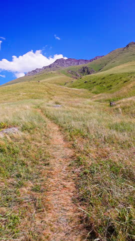 Winding unpaved path invites exploration through grassy hillside towards rocky mountain peak. Scenic view under clear sky evokes tranquility and adventure.