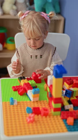 Little girl holding toy blocks with hands at a play table in a building surrounded by stuffed animals and a plastic bin; quiet concentration.