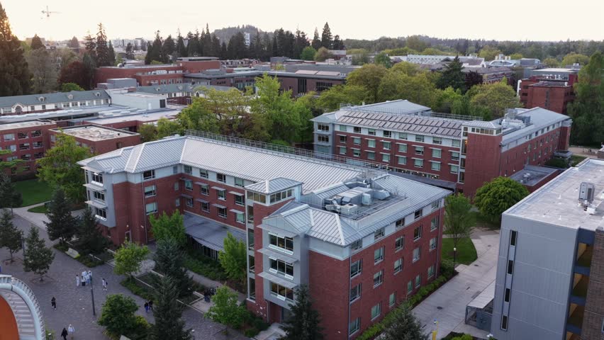 Red brick university campus buildings with metal rooftops, tree-lined walkways, students on paths, and forested hillside under soft evening light. Eugene, Oregon, USA. Aerial flyover shot. Quiet scene