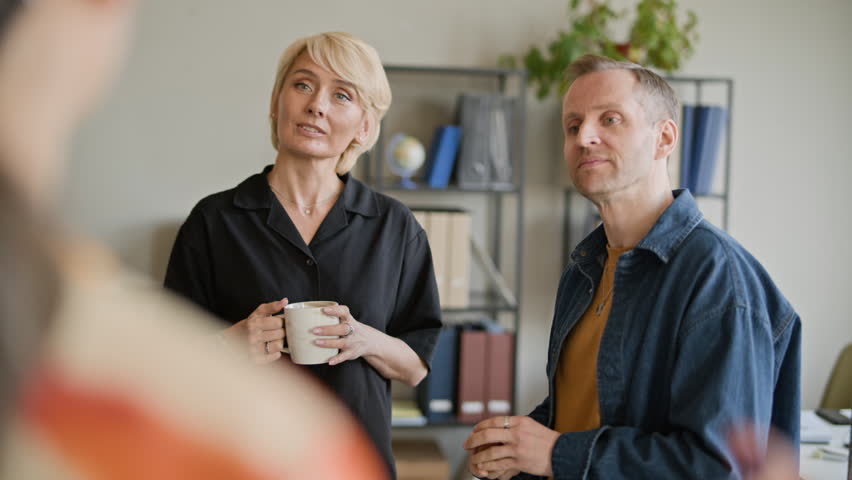 Over the shoulder view of woman talking to two adult Caucasian colleagues while having coffee break in office