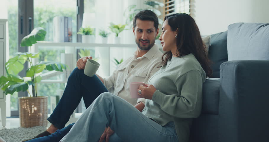 Happy young adult Hispanic couple drinking coffee and relaxing sit on floor in living room at home. Domestic lifestyle, positive weekend leisure, cheerful relationship bonding, family love concept.