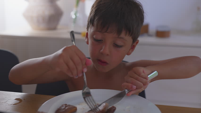 Boy eating meat with fork and knife, leaning forward and focused on cutting food, showing independence and developing motor skills during indoor mealtime