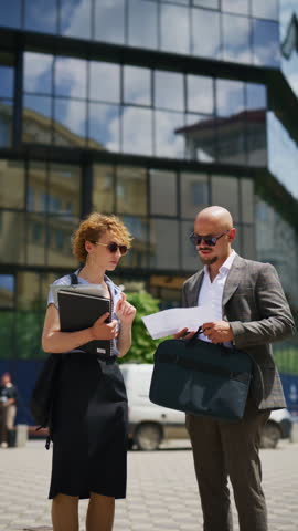 Two professional business colleagues discuss documents and collaborate outdoors. A man and woman review paperwork in front of a modern office building.