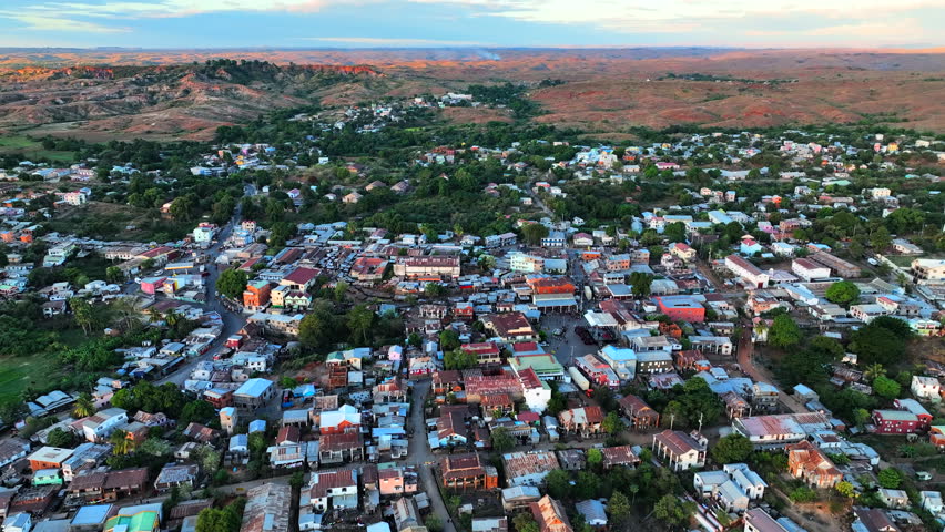 Low aerial shot over the rooftops of Antsirabe, Madagascar, at dusk. Terracotta and corrugated tin rooftops fill the frame as the golden-hour light warms the highland town surrounded by distant red hills.
