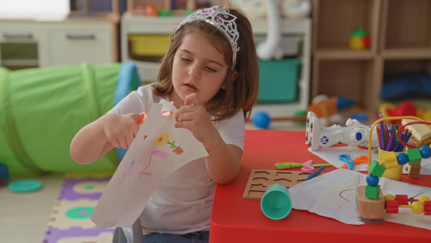 Girl wearing tiara cuts paper with scissors and draws with crayon at a small red table in building, holding the paper with her hands; concentration.
