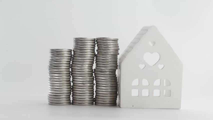 A hand reaches out and puts coins in a stack near a small white ceramic house on a white table. The stack grows noticeably as coins are added one after another,