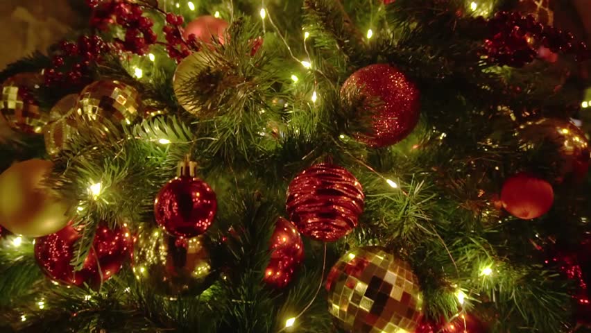 Close up view of decorated Christmas tree. Numerous glowing warm white string lights illuminate textured red and shimmering gold sphere ornaments. Artificial pine branches provide festive holiday back