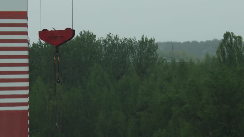 Close up view of heavy duty red crane hook and chain assembly, part of tall red and white striped crane mast, towering above dense green forest under flat gray overcast sky.