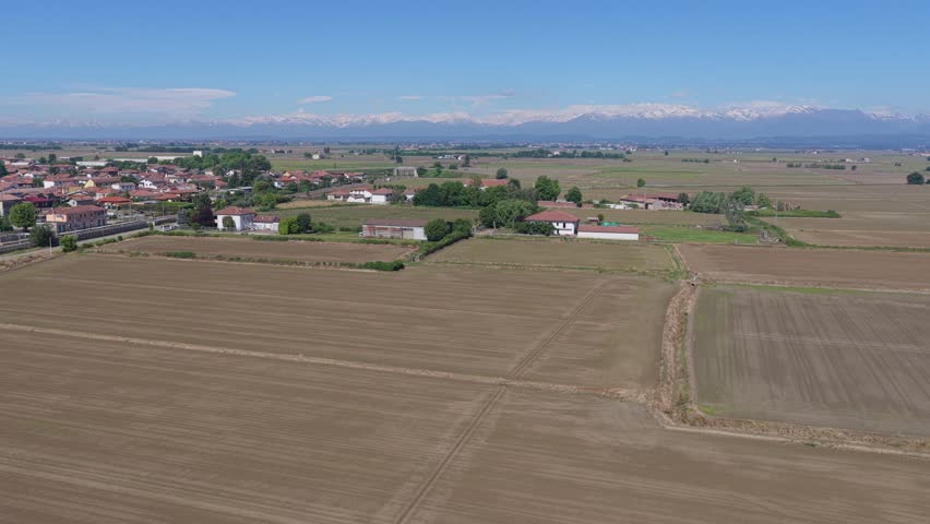 View of plowed agricultural fields and a small village with snow-capped Alps in the background under a clear blue sky San Germano Vercellese, Piedmont, Italy.