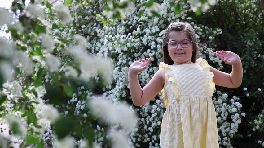 Happy little girl with glasses relaxing and holding hair behind head near blossoming white spirea bushes
