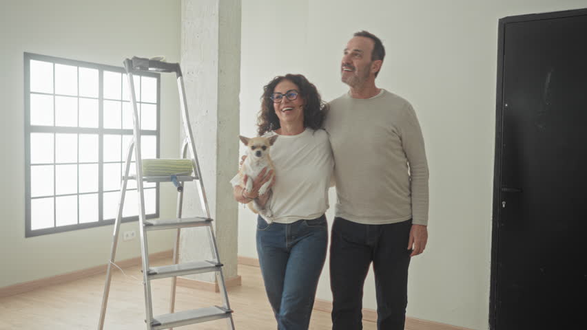 Man and woman embrace while holding a small dog beside a step ladder in building; new home contentment.