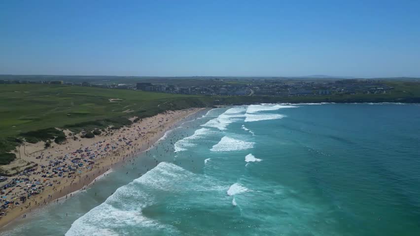 Newquay, North Cornwall, England: DRONE VIEWS: The drone flies over rolling waves heading towards Fistral Beach; in the sea, surfers wait to catch a wave as sunbathers relax on the beach.
Newquay is a civil parish, seaside resort, a regional centre for aerospace industries with an airport, a spaceport and a fishing port on the North Atlantic coast; it is located approximately 12 miles north of Truro and 20 miles west of Bodmin.
The town is bounded to the south by the River Gannel and its associated salt marsh, and to the north-east by the Porth Valley. The western edge of the town meets the Atlantic at Fistral Bay.
