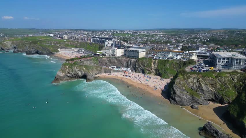 Newquay, North Cornwall, England: DRONE VIEWS: The drone flies towards Great Western Beach showing a general view of the town of Newquay and Tolcarne Beach to the left of picture; the drone camera tilts down to give an overhead shot of surfers in the sea and sunbathers on Great Western Beach.
Newquay is a civil parish, seaside resort, a regional centre for aerospace industries with an airport, a spaceport and a fishing port on the North Atlantic coast; it is located approximately 12 miles north of Truro and 20 miles west of Bodmin.
The town is bounded to the south by the River Gannel and its associated salt marsh, and to the north-east by the Porth Valley. The western edge of the town meets the Atlantic at Fistral Bay.