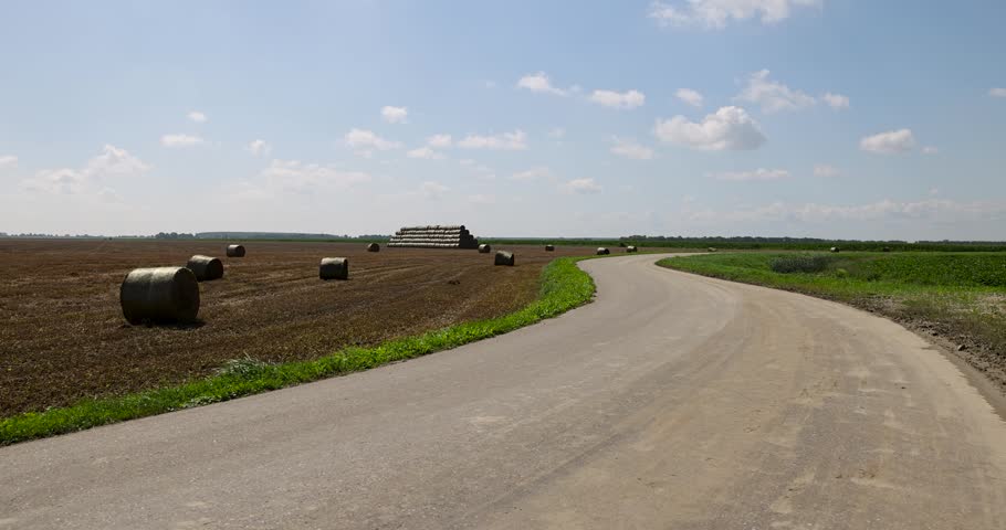 A bright empty road, this winding road divides a cleared area with rolls of straw and a bright green meadow on a clear summer day.