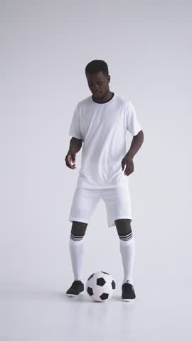 Young male football player dressed in white sportswear skillfully controlling and juggling a classic black and white soccer ball on a plain background during practice