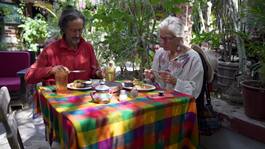 Mature woman shares a meal with a local man under greenery in Mulege Baja California Sur Mexico while enjoying the local culture.