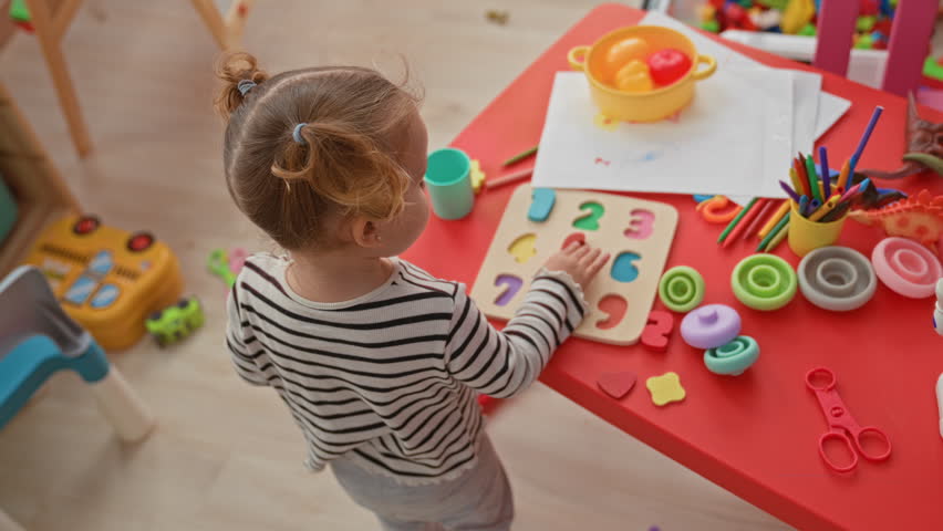 Girl blonde toddler placing a green puzzle piece with her hand on a red table beside stacking rings and crayons in a playroom building; concentration learning.