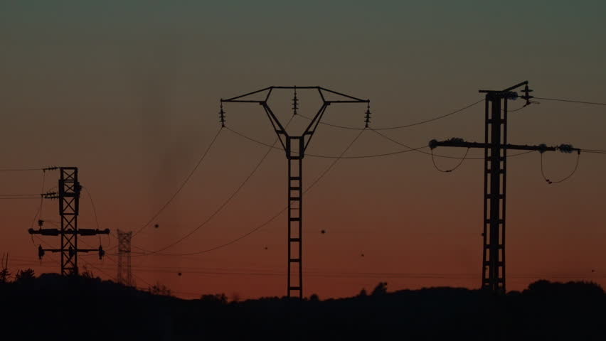 High voltage electricity pylon cast dark silhouette against vivid red and orange gradient sunset sky over a rural landscape
