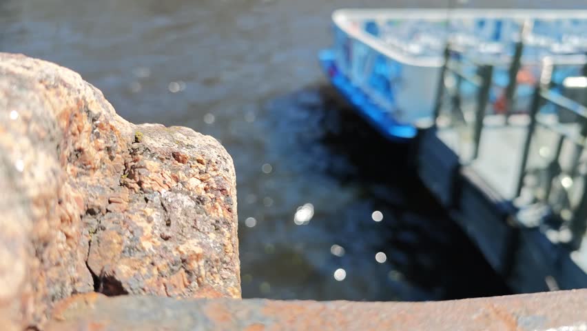 Close-up view of weathered metal railing edge overlooking a river with a boat docked nearby, sunlight reflecting off the water surface in a serene outdoor setting